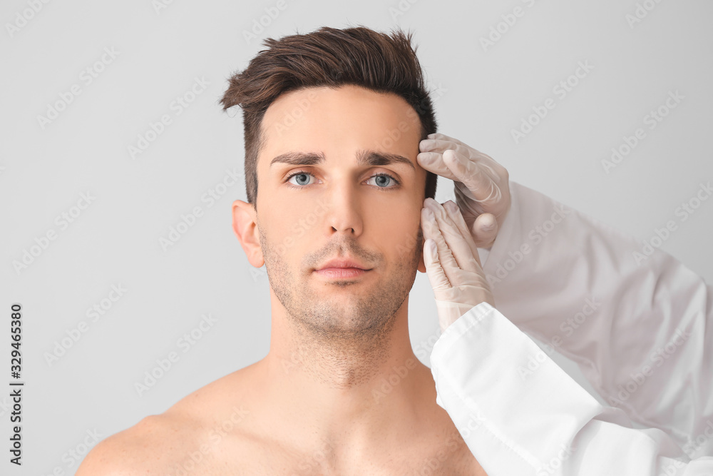 Plastic surgeon touching face of young man on light background