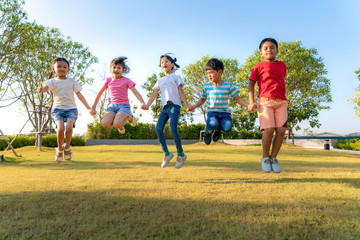 Wall Mural - Large group of happy Asian smiling kindergarten kids friends holding hands playing and jumping together during a sunny day in casual clothes at city park.