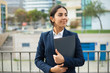 © Mangostar - Happy businesswoman holding folder. Smiling young businesswoman in formal wear holding papers and looking aside on street. Professional occupation concept