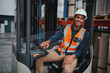 © StratfordProductions - Portrait of happy young african forklift manager wearing safety vest and white hardhat transporting goods from one shelf to another while looking in camera in warehouse