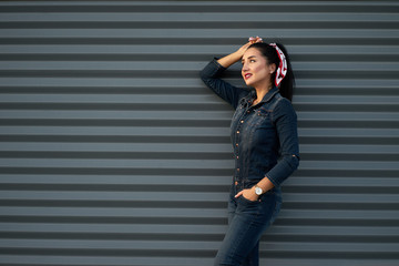 New trendy collection. Portrait pretty young fashionable brunette girl with long hair, charming smile and red painted lips wearing modern hipster denim overalls posing on grey urban wall background.