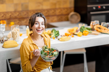 Wall Mural - Young and cheerful woman eating salad at the table full of healthy raw vegetables and fruits on the kitchen at home. Concept of vegetarianism, healthy eating and wellness