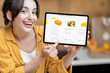 © rh2010 - Woman holding digital tablet with launched online shopping market while standing on the kitchen with lots of fresh food on the table