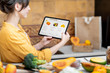 © rh2010 - Woman holding digital tablet with launched online shopping market while standing on the kitchen with lots of fresh food on the table