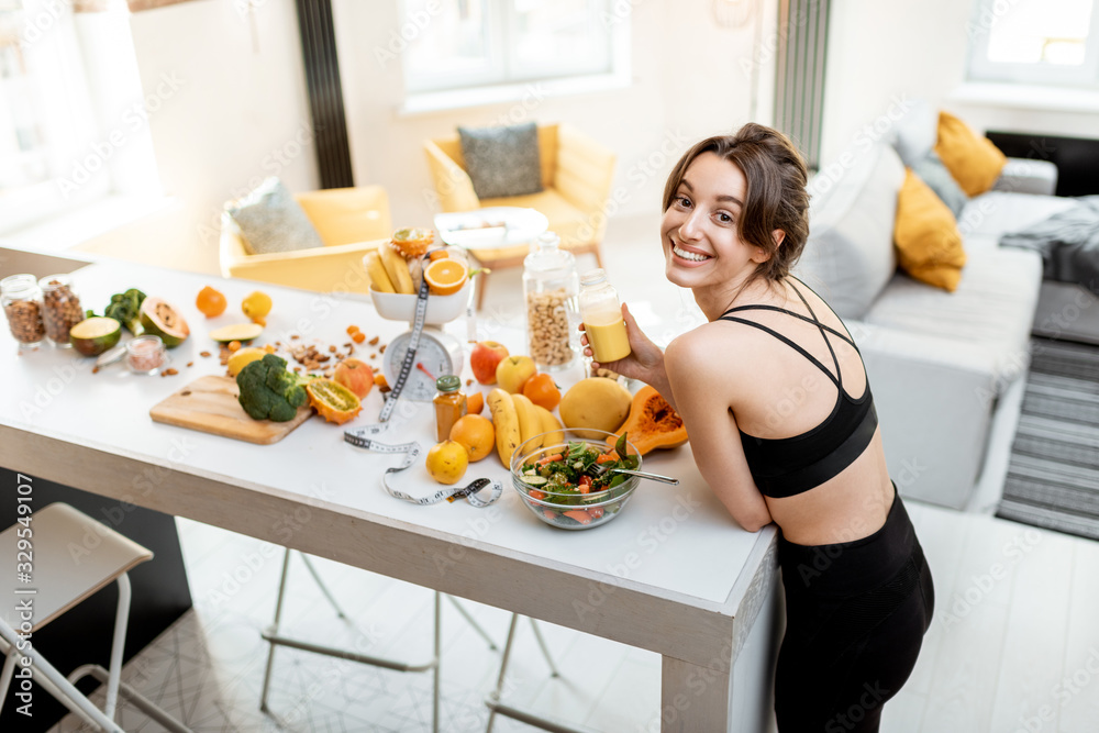 Portrait of an athletic woman having a break, drinking juice while standing with lots of healthy fresh food on the kitchen. Concept of losing weight, sports and healthy eating - obrazy, fototapety, plakaty 