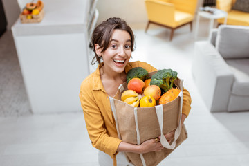 Portrait of a young and cheerful woman standing with shopping bag full of fresh fruits and vegetables at home. Concept of online shopping and healthy eating