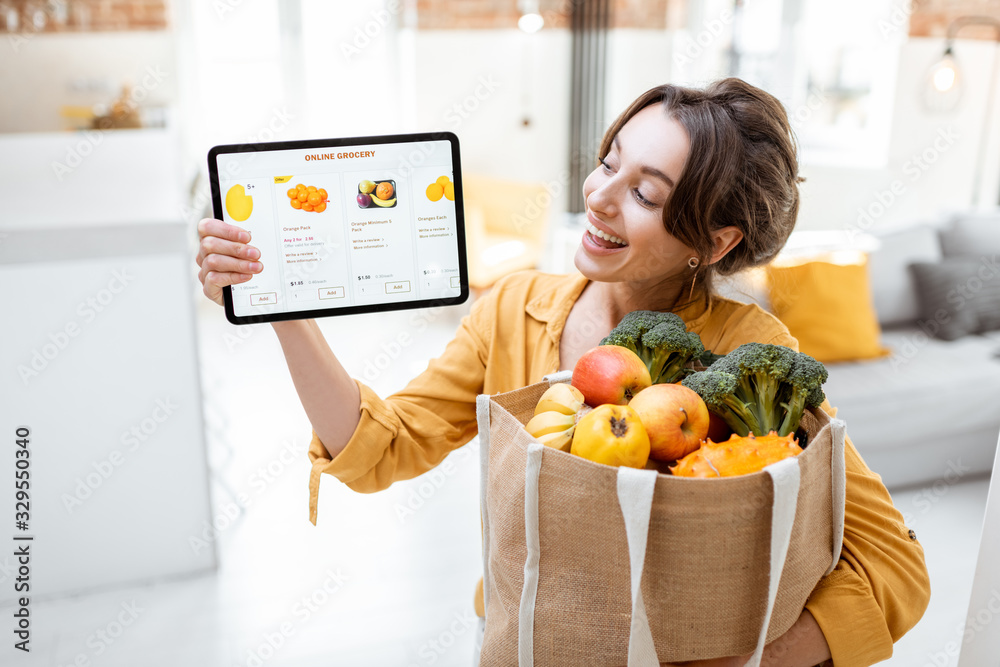 Happy woman holding a digital tablet with launched online store while standing with shopping bag full of fresh products at home. Concept of buying online using mobile devices - obrazy, fototapety, plakaty 