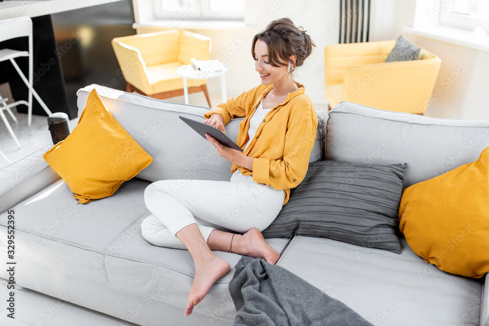 Young and cheerful woman using a digital tablet while sitting relaxed on the couch at home. Concept of a leisure activities with mobile devices at home - obrazy, fototapety, plakaty 