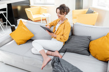 Young And Cheerful Woman Using A Digital Tablet While Sitting Relaxed On The Couch At Home. Concept Of A Leisure Activities With Mobile Devices At Home