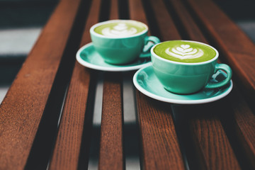 Wall Mural - Two cups of cappuccino with latte art on wooden background. Beautiful foam, greenery ceramic cups, stylish toning, place for text.