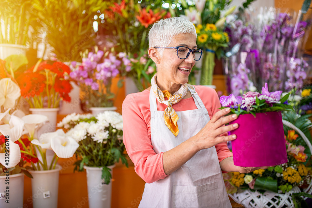 Senior woman sales flowers in flower shop. Happy smiling mature florist woman having eyeglasses and working at flower shop. Portrait of successful modern florist wearing apron. - obrazy, fototapety, plakaty 