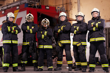 Wall Mural - Six professional firefighters posing together. Firefighters wearing uniforms and protective helmets. Firetruck in the background.