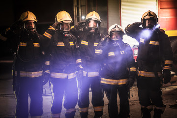 Wall Mural - Group of professional firefighters posing. Firemen wearing uniforms, protective helmets and oxygen masks. Smoke and firetrucks in the background.