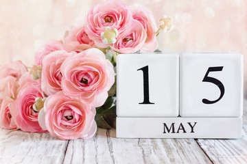 White wood calendar blocks with the date May 15th. Selective focus with pink ranunculus in the background over a wooden table.