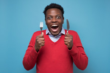 Young Hungry African Man Holding A Fork And A Knife Waiting For Delicious Meal. Isolated On Blue Background
