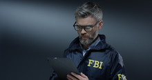 Good Looking Caucasian Grey-haired Male FBI Policeman In Glasses Tapping And Scrolling On The Tablet Device While Browsing.