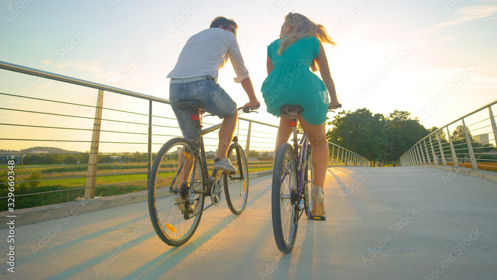LOW ANGLE: Unrecognizable couple riding their bikes across an overpass at sunset - obrazy, fototapety, plakaty 