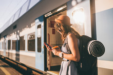 Railroad Theme. Beautiful Young Woman With A Backpack Uses The Phone While Standing Near The Railroad Train On The Platform. Cheap Travel