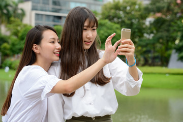 Two happy young beautiful Asian teenage girls taking selfie together at the park