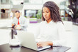 © Mangostar - Confident young businesswoman using computer in co-working space. African American business woman looking at laptop screen, man using tablet in background. Young businesswoman concept
