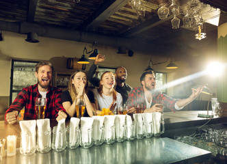 Wall Mural - Sport fans cheering at bar, pub and drinking beer while championship, competition is going. Multiethnic group of friends excited watching translation. Human emotions, expression, supporting concept.