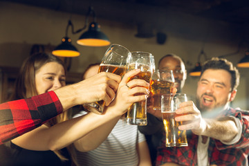 Wall Mural - Sport fans cheering at bar, pub. Clinking beer glasses while watching championship, competition. Multiethnic group of friends excited in translation. Human emotions, expression, supporting concept.