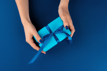 Wall Mural - Woman holding gift box with ribbon against classic blue background, top view