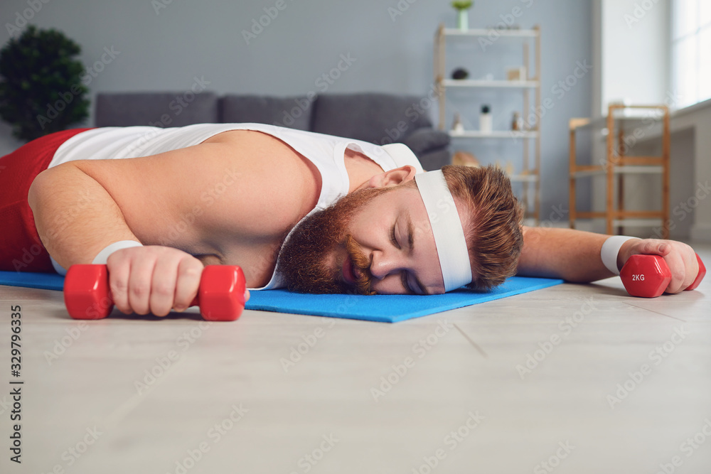 A lazy fat man lies with dumbbells on the floor of the house. - obrazy, fototapety, plakaty 