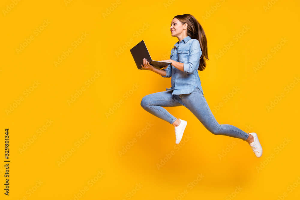 Full body profile photo of pretty business lady jump high holding notebook hands hurry work browsing laptop wear casual denim outfit white sneakers isolated yellow color background - obrazy, fototapety, plakaty 