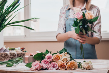 Hands of young woman florist working with fresh flowers making bouquet of pink roses on table.