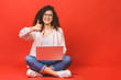 Happy young curly beautiful woman sitting on the floor with crossed legs and using laptop on red background. Thumbs up.