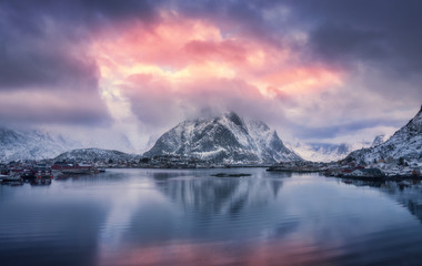 Wall Mural - Aerial view of snowy mountain, village on sea coast, blue sky with red clouds at sunset in winter. Top view of Reine, Lofoten islands, Norway. Landscape with rocks, houses, rorbu, reflection in water