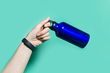 Close-up of female hand holding a reusable eco, steel thermo water bottle of phantom blue color, isolated on aqua menthe background.