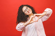 Portrait of a smiling young curly woman showing heart gesture with two hands and looking at camera isolated over red background.