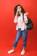 © denis_vermenko - Full length portrait of a female student walking and looking at a mobile phone isolated on red background.
