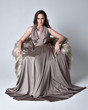 Portrait of a pretty brunette girl wearing a long silver evening gown, full length seated pose against a studio background.