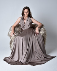 Canvas Print - Portrait of a pretty brunette girl wearing a long silver evening gown, full length seated pose against a studio background.