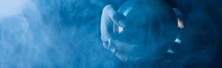 Wall Mural - Cropped view of female hands with crystal ball and smoke around on dark blue background, panoramic shot