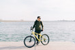 © Дарья Фомина - a young man in khaki sportswear and glasses stands on a pontoon near the lake and holds a yellow Bicycle near him on a Sunny spring day