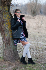 Melancholy, very beautiful woman in glasses and a black jacket posing near a tree