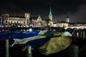 Night time view of Zurich old town from the Limmat river side covered up small boats on pier and illuminated church towers in the background