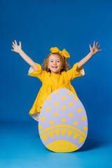 Wall Mural - child a blonde girl in a yellow dress is smiling standing with a row of large decorative Easter eggs on a blue background