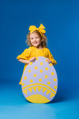 Wall Mural - child a blonde girl in a yellow dress is smiling standing with a row of large decorative Easter eggs on a blue background