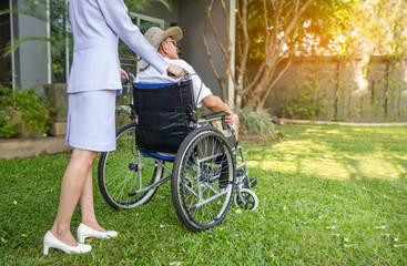 Young nurse helping elderly man on wheelchair outdoor in garden.