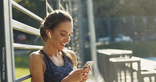 Cuadros en Lienzo Pretty smiled Caucasian woman in sporty style and airpods tapping and texting message on smartphone on sunny warm day