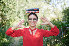 Young Blond Woman, Wearing Red Shirt And Eyeglasses, Holding Two Books On Her Head. Student With Black And Red Books, Smiling. Studying Fun. Pretty Girl, Balancing With Books On Her Head,