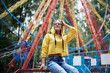 canvas print picture - Young blond woman, wearing yellow hoody and blue jeans, spending time in amusement theme park in summer. Three-quarter portrait of girl, sitting on railing in front of colorful ferris wheel, smiling.