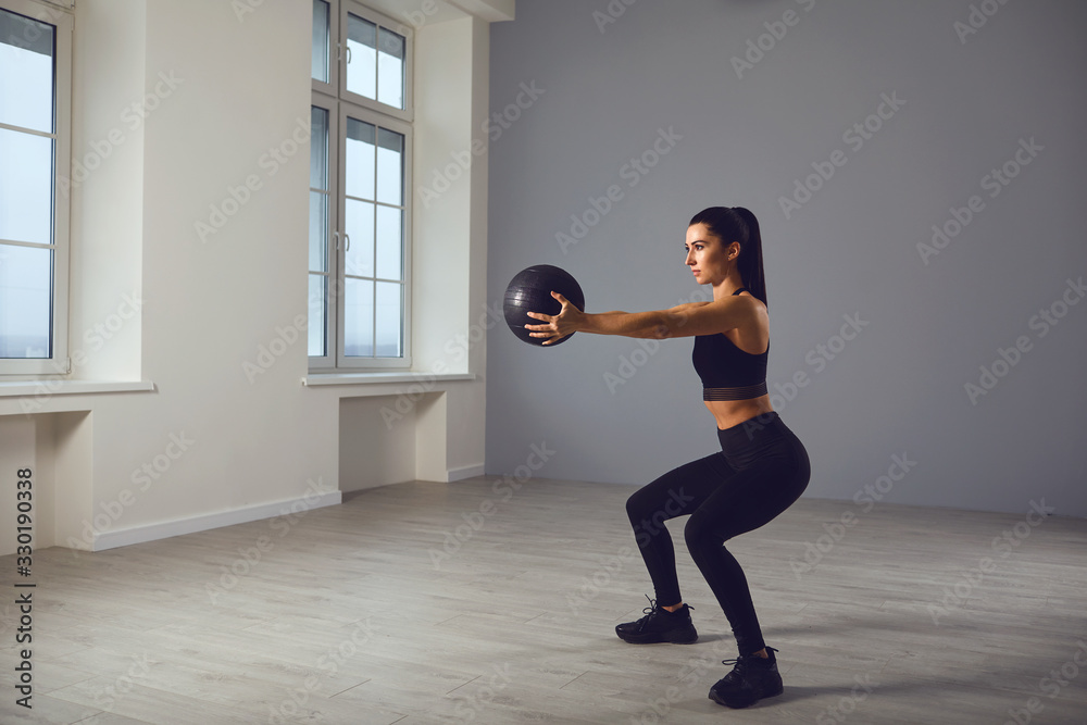 The girl goes in for sports with a medical ball. A sports girl does exercises in a room indoors. - obrazy, fototapety, plakaty 