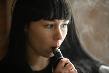 Wall Mural - Vaping teenager. Young pretty white caucasian teenage brunette girl with blue eyes and problem skin smoking an electronic cigarette indoors. Deadly bad habit. Vape activity.