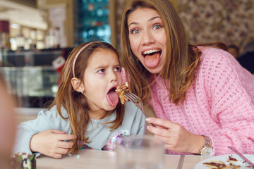 Portrait of small caucasian girl with mouth opened sitting by the table at restaurant while her mother is feeding brunette daughter holding fork and food in day posing looking to the camera